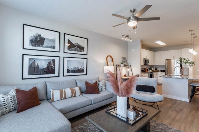 Spacious furnished living room with an open layout, featuring a ceiling fan and a clear view of the kitchen in the background, located at Destination at Arista apartments.